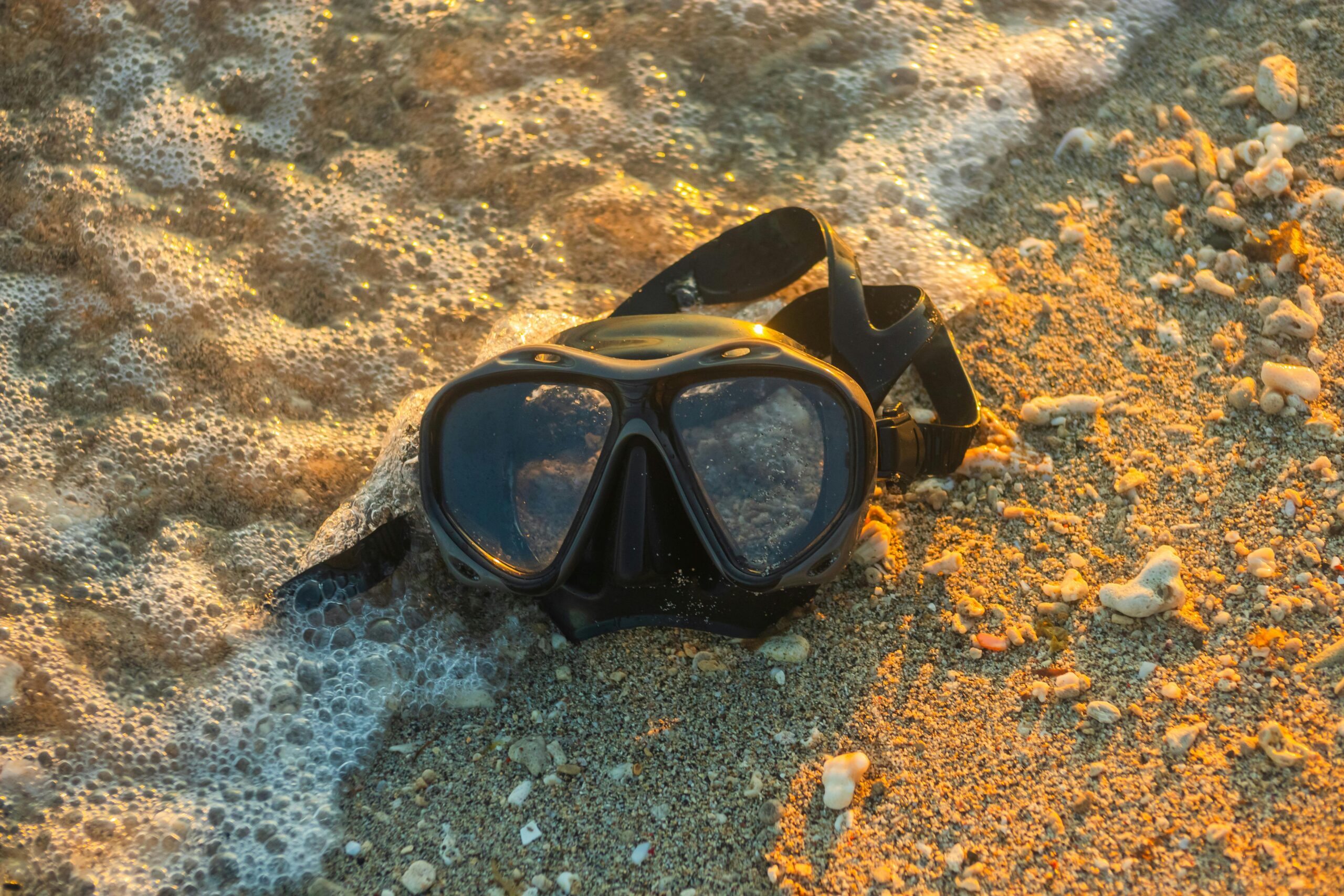 A scuba mask rests on a sandy shoreline as waves gently wash over it during sunset.